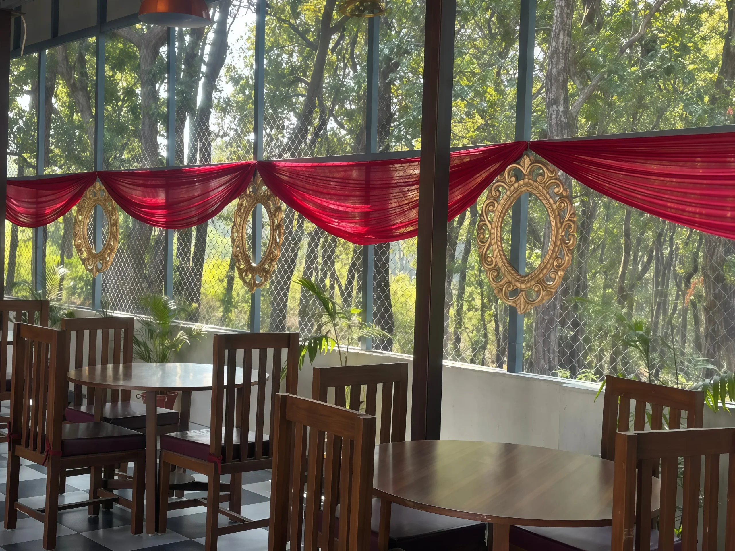Sunlit restaurant interior featuring wooden tables and chairs beside large glass windows decorated with red draped fabric and ornate gold frames, overlooking a green outdoor landscape with trees.